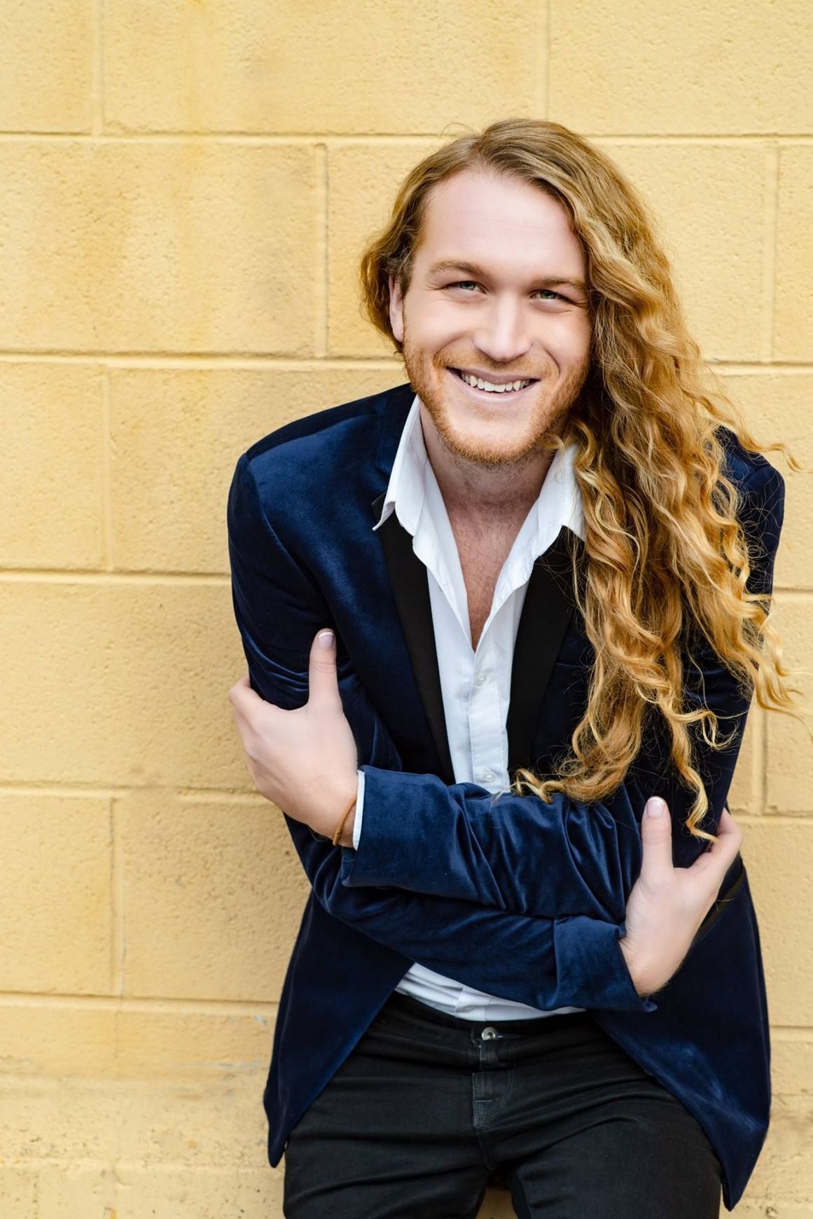 A man with long hair is sitting in front of a brick wall.