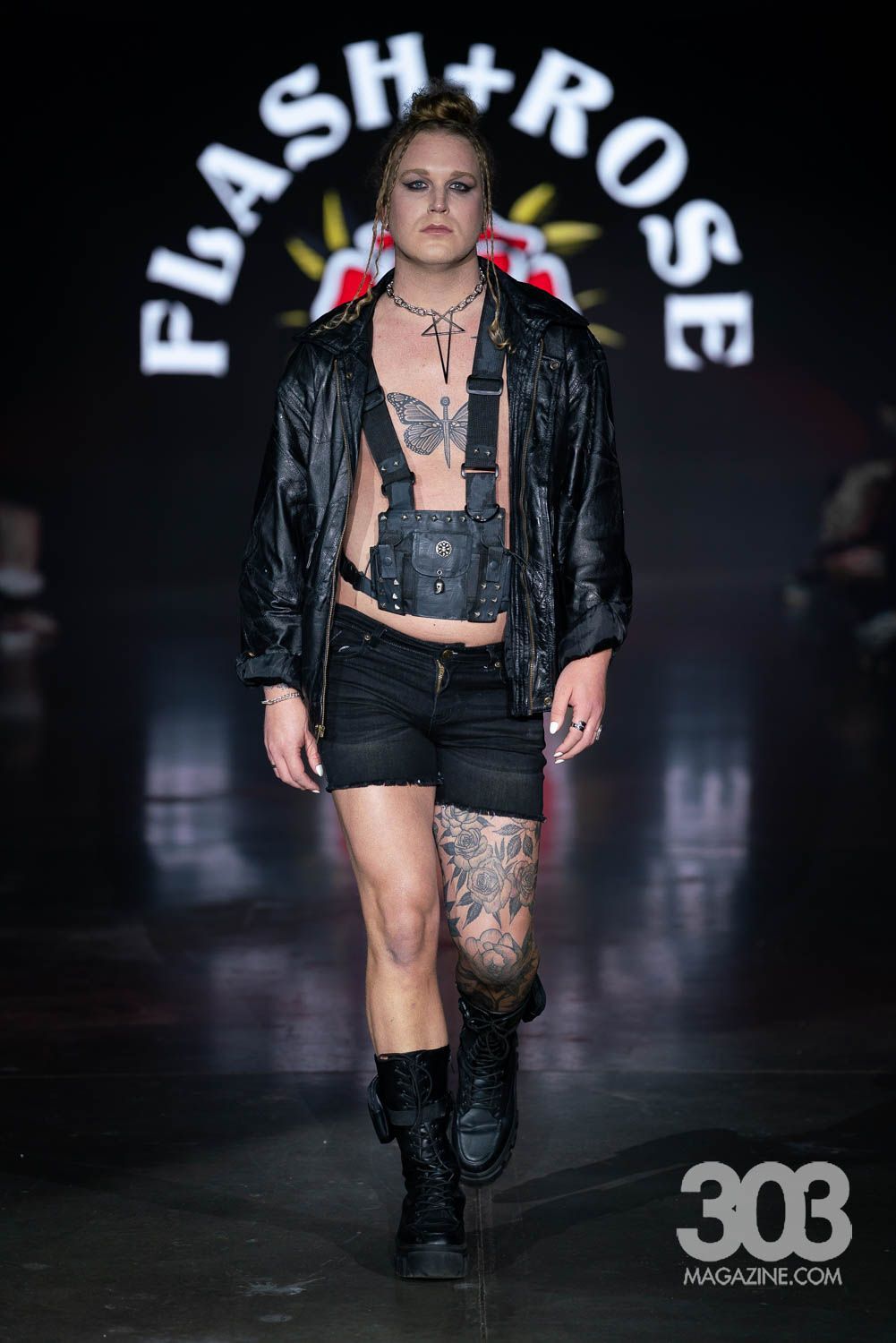 A man is walking down a runway wearing a leather jacket and shorts.