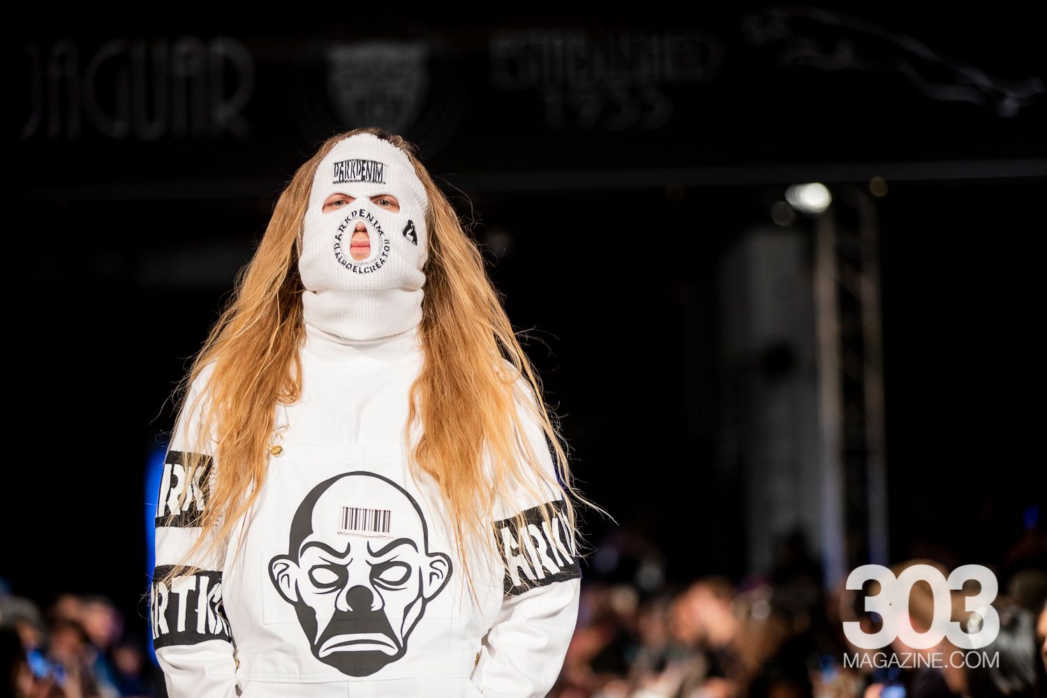 A woman is walking down a runway wearing a white hoodie with a mask on her face.