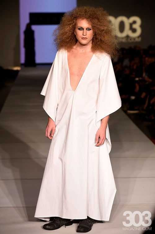 A man with red hair is walking down a fashion show runway wearing a white dress.