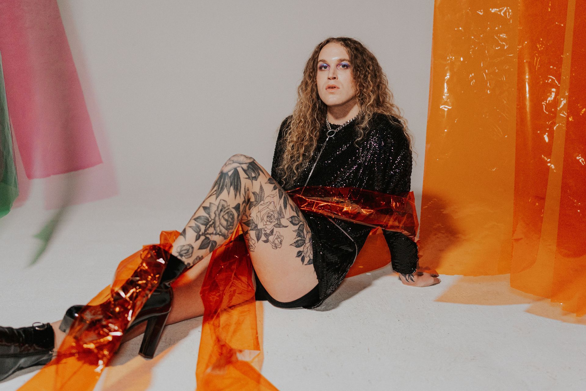A woman with tattoos is sitting on the floor in front of an orange curtain for a photo shoot.