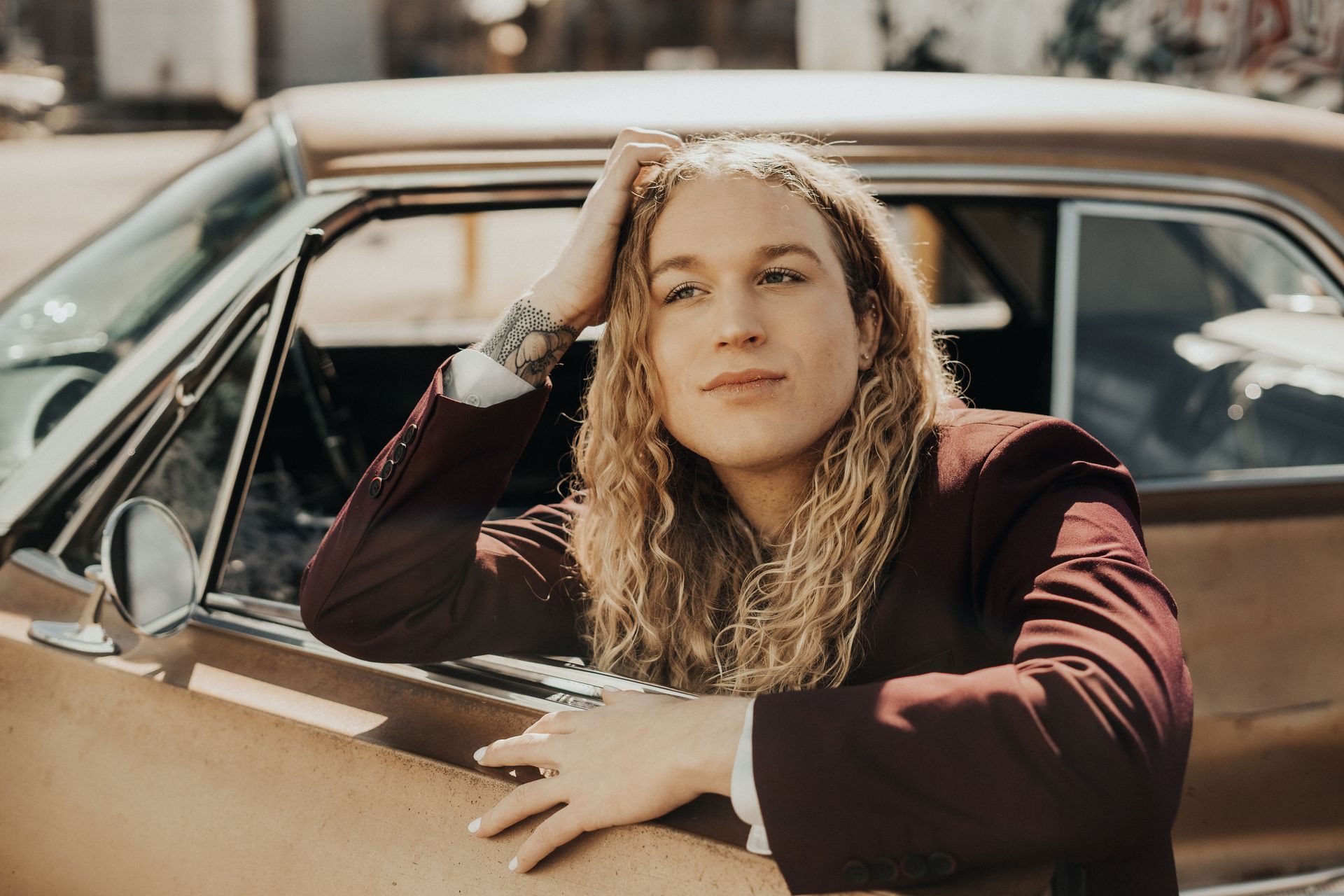 A man with long hair is leaning on the side of a car for a photo shoot.