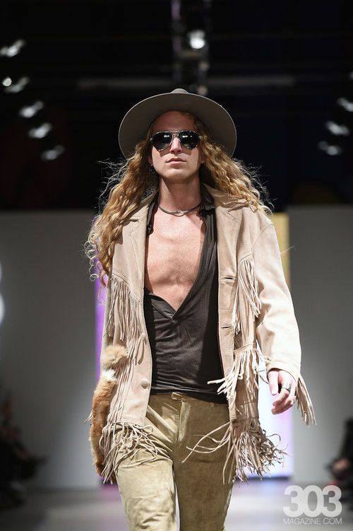 A man with long hair is walking down a runway wearing a hat and sunglasses.