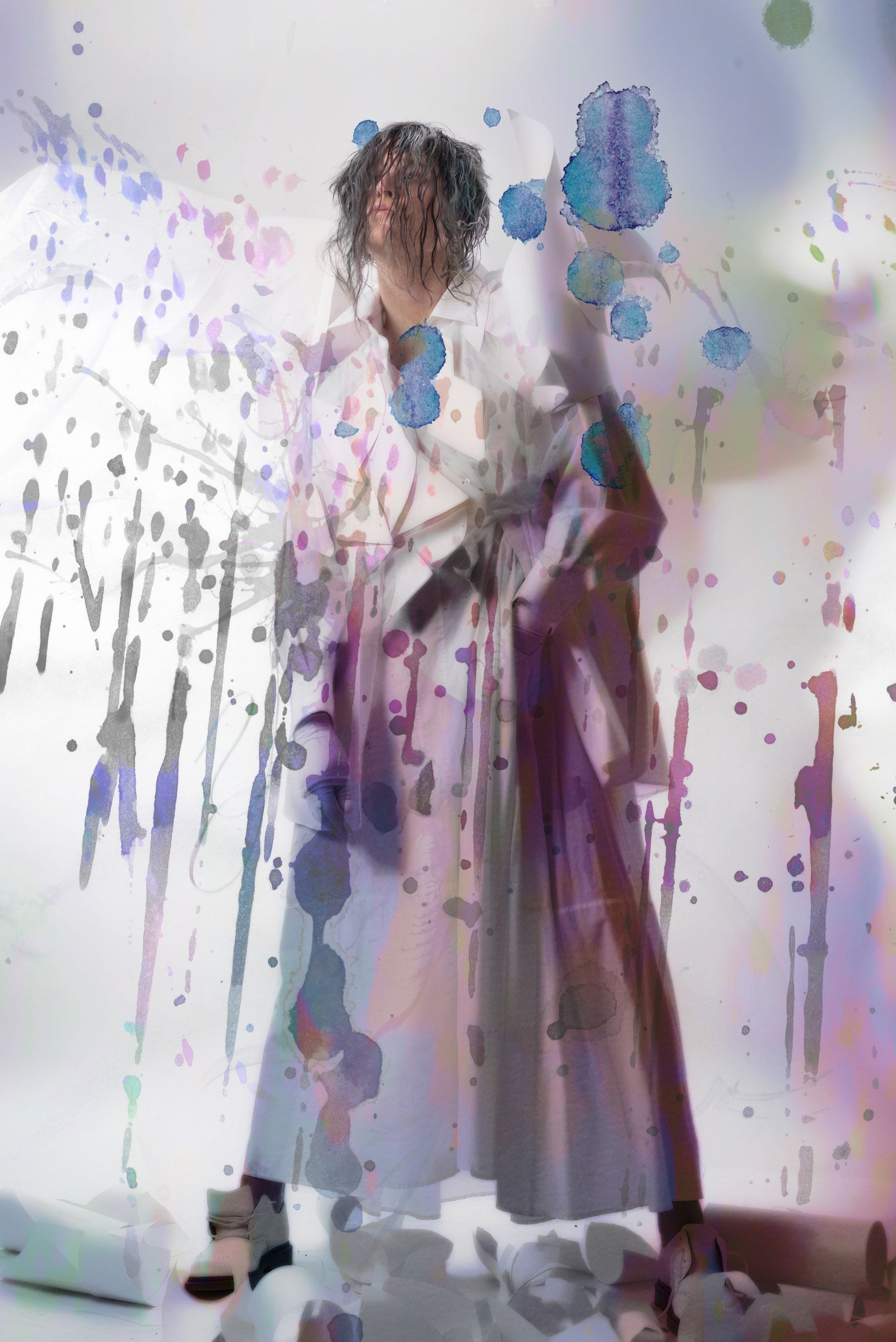Scotty in a long dress is standing in front of a wall covered in paint for a fashion magazine photo shoot.