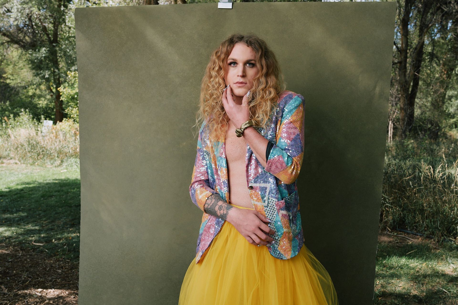 Scotty in a colorful jacket and yellow skirt is standing in front of a green wall for a fashion photo shoot.