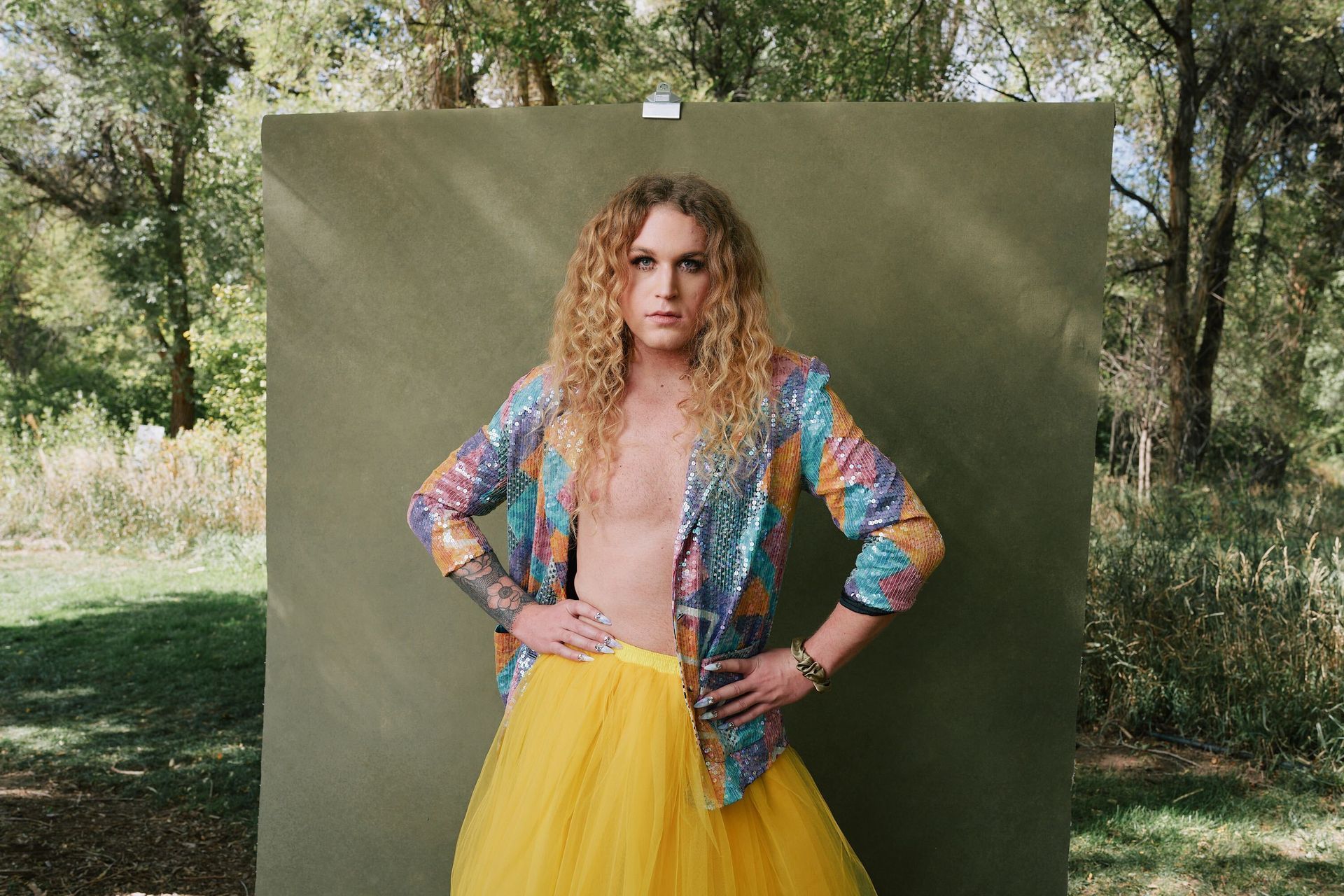 Scotty in a yellow skirt and a colorful jacket standing in front of a green wall for a fashion photo shoot.
