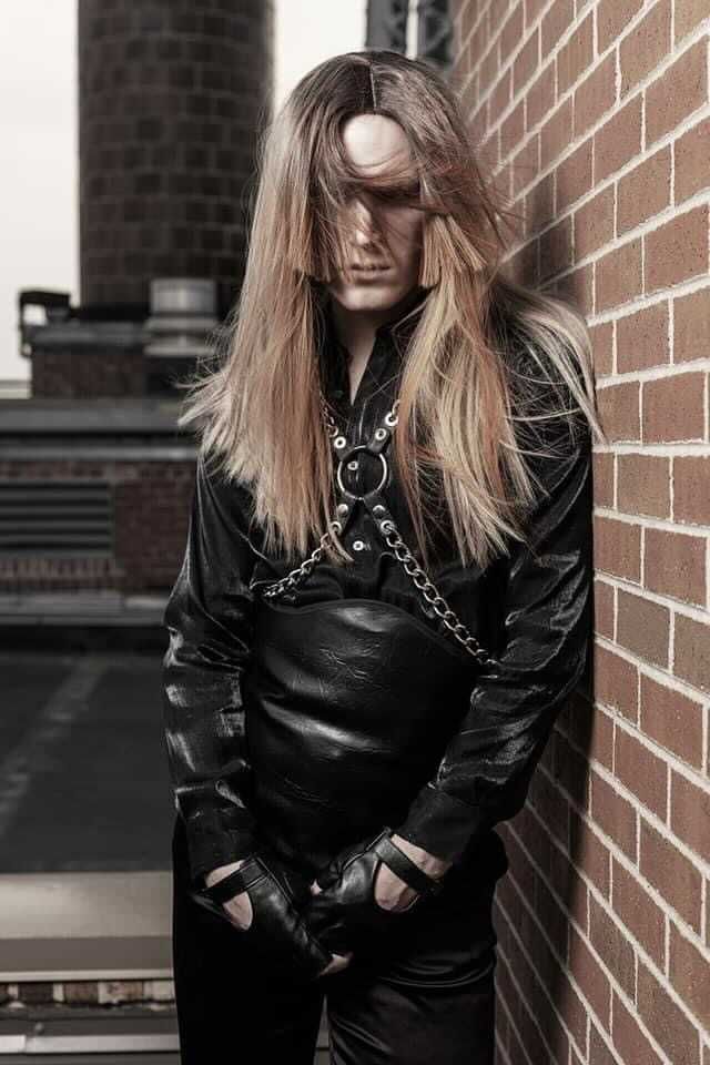 A young man with long hair is leaning against a brick wall for a photo shoot.