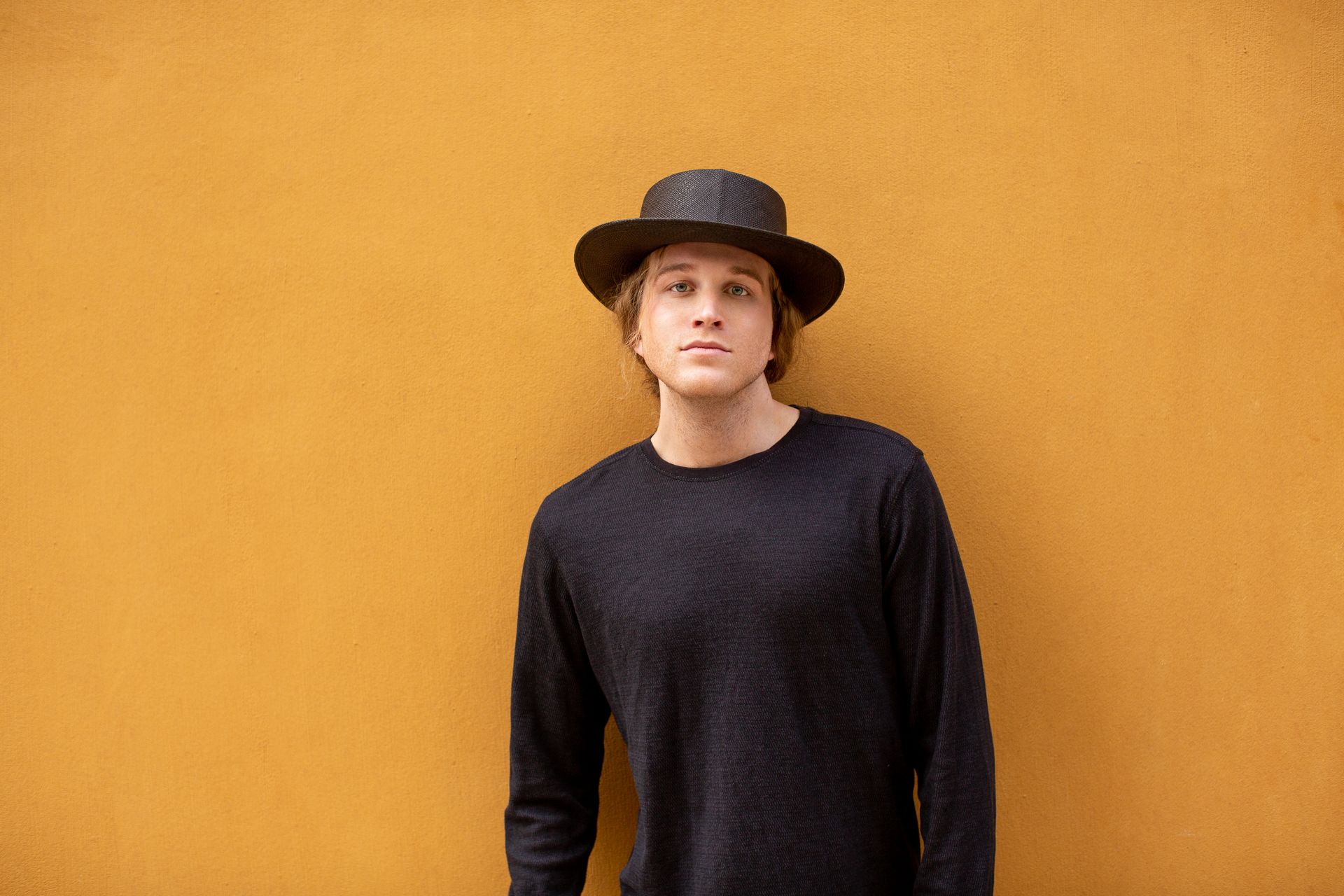 A young man wearing a hat and a black sweater is standing in front of a yellow wall.
