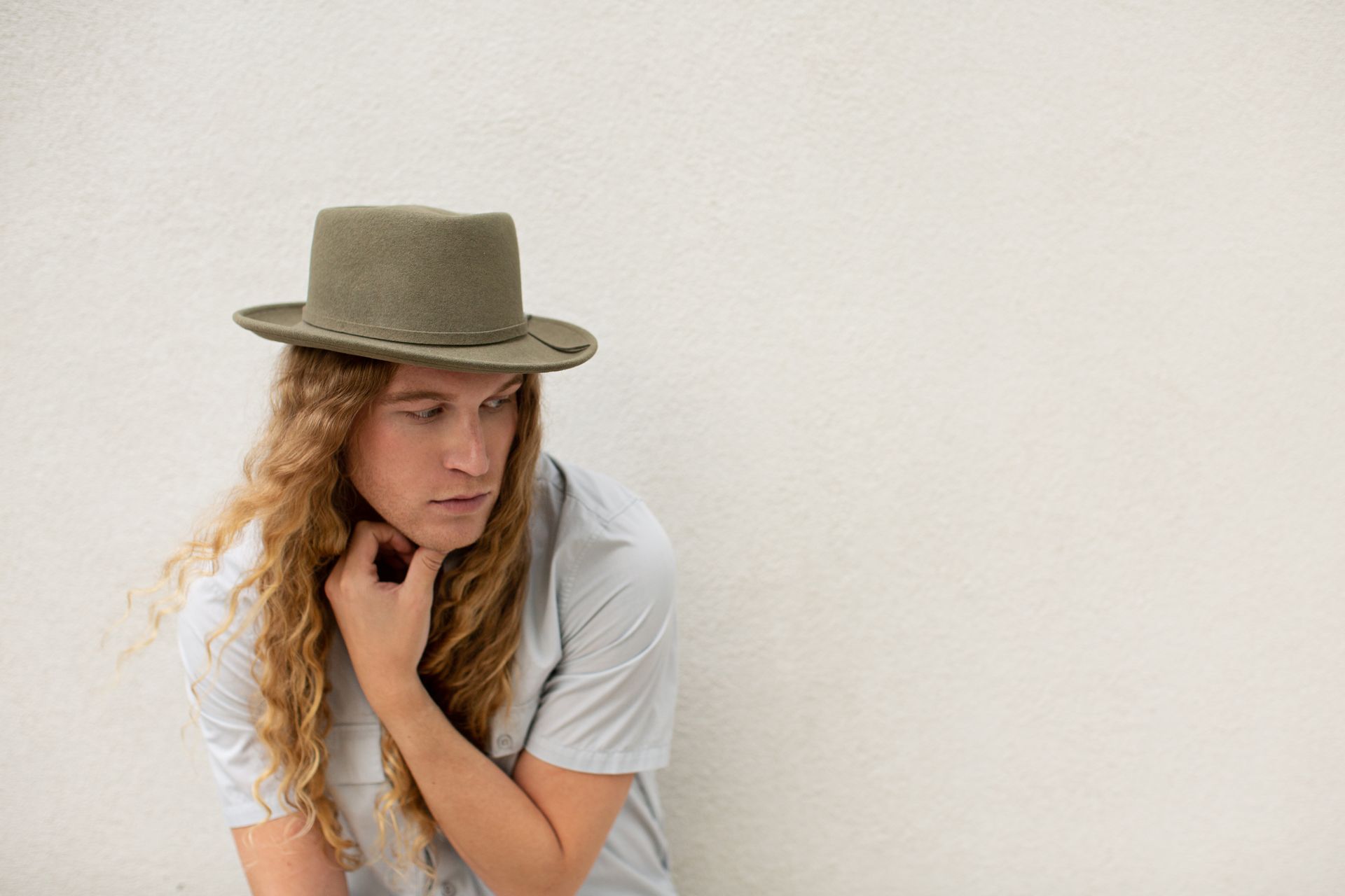 A young man with long hair wearing a hat is sitting in front of a white wall for a fashion photo shoot.