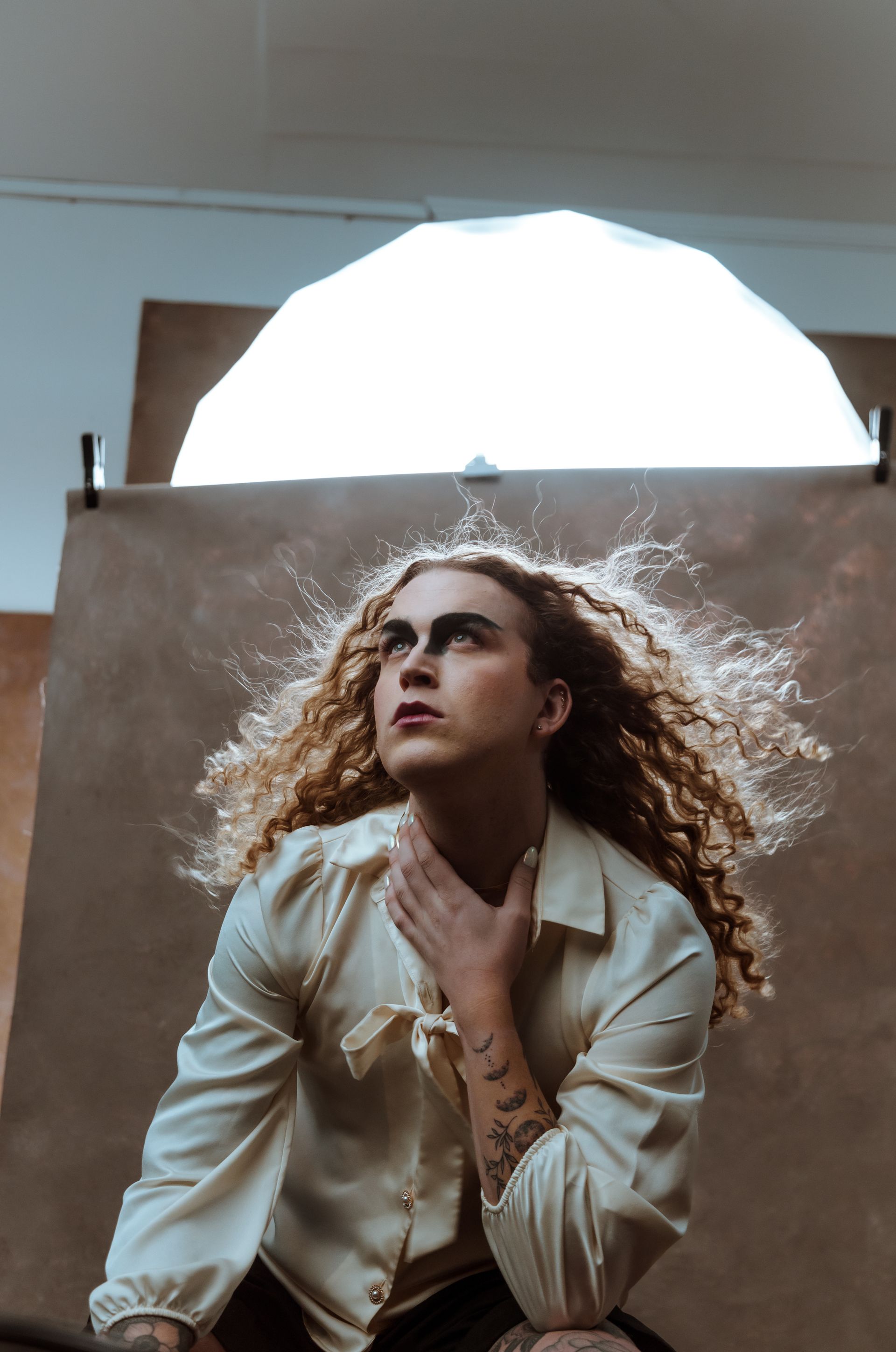 A man with long curly hair is sitting in front of a light for a fashion photo shoot.