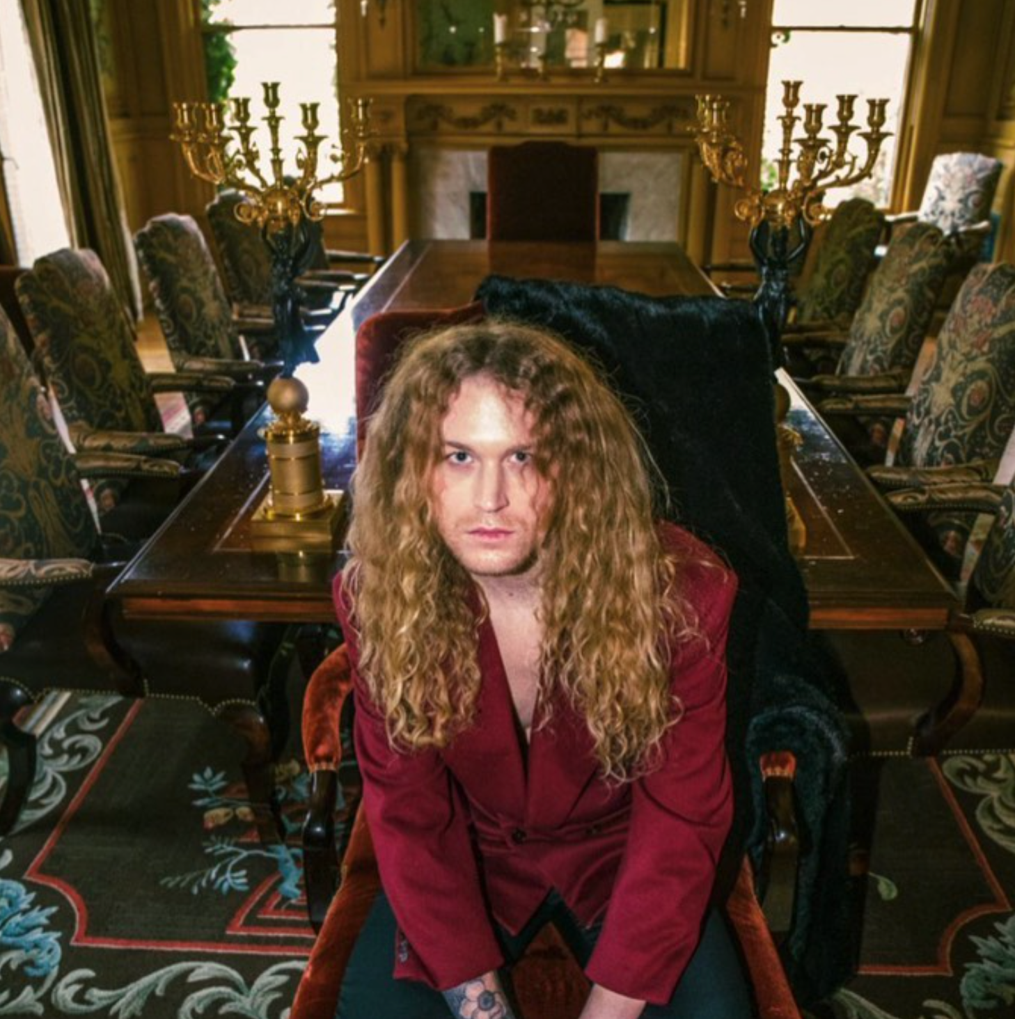 A man with long hair is sitting in a chair in front of a long table for a photo shoot for a fashion magazine.