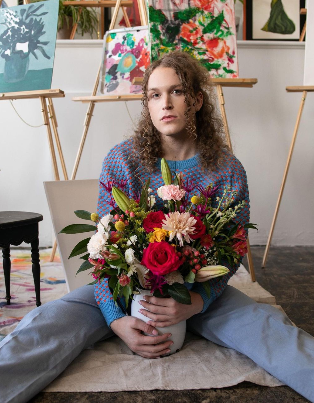 A woman is sitting on the floor holding a vase of flowers for a fashion photo shoot.