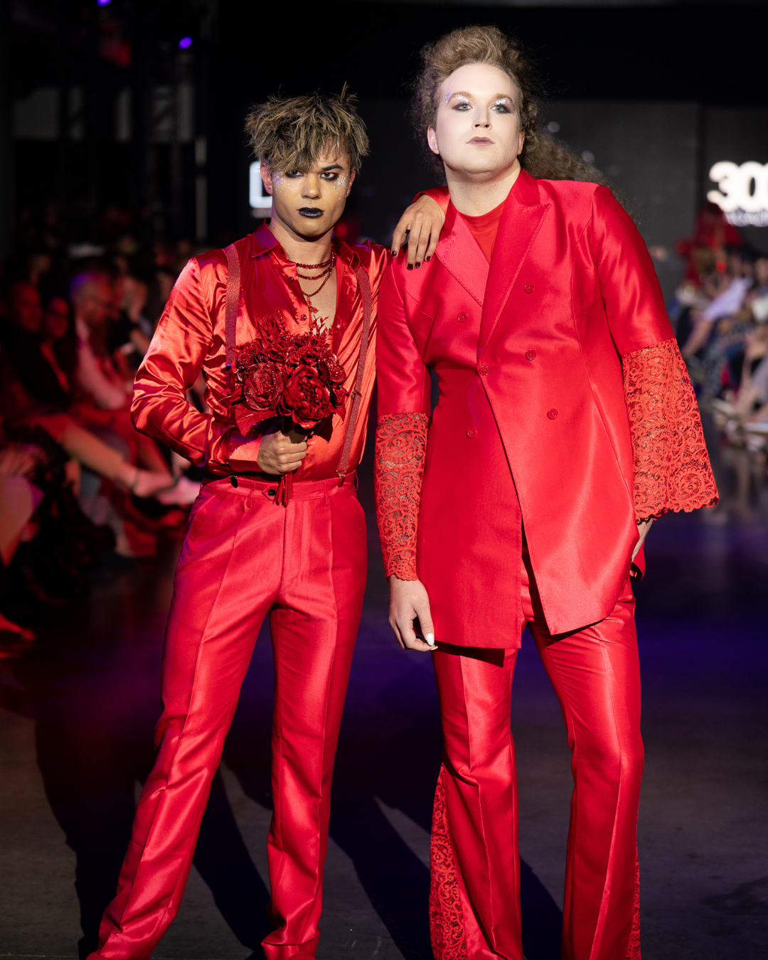 Two women in red suits are standing next to each other on a runway for Denver Fashion Week.