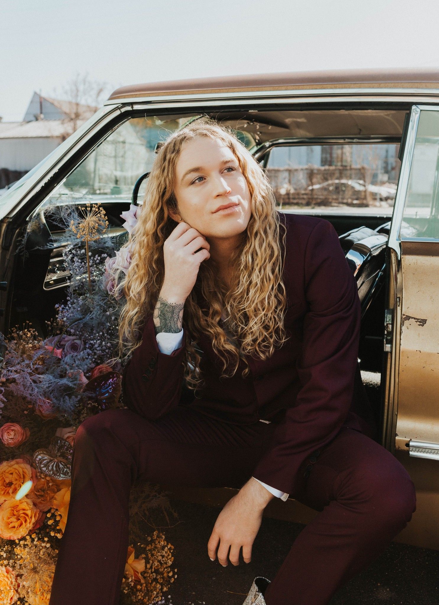 Scotty wearing a maroon suit posing in a car with flowers, modeling for a photoshoot