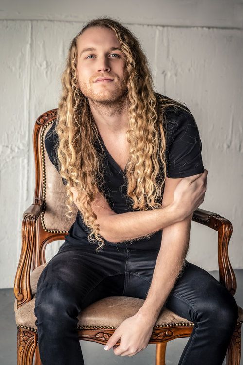 A man with long blonde hair is sitting in a chair with his arms crossed for a fashion photo shoot.