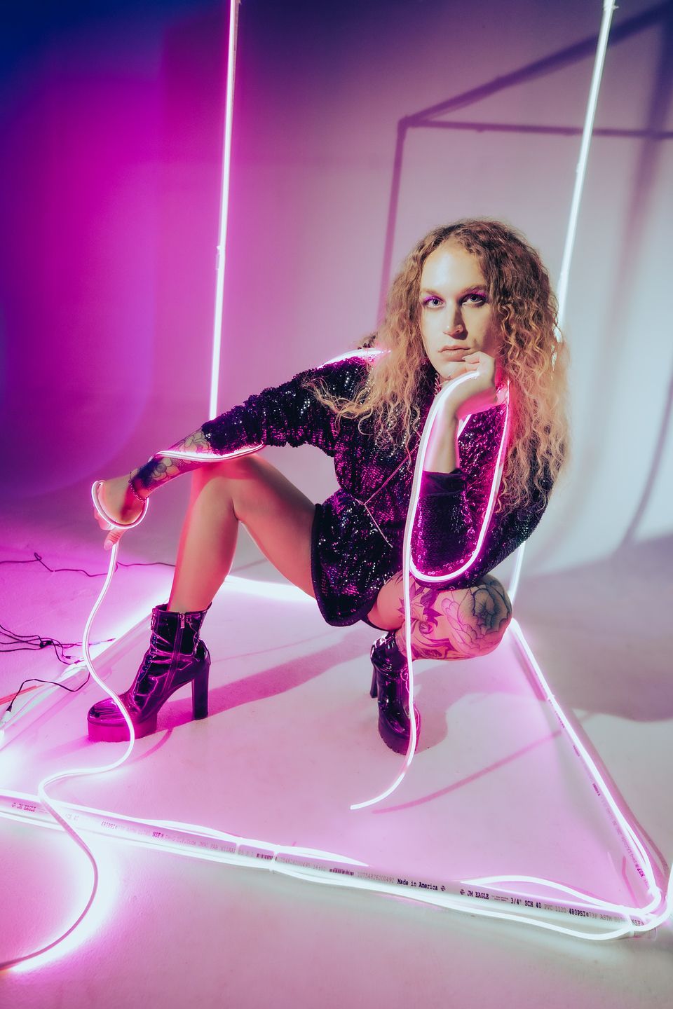 Scotty in a model pose for a photoshoot in a black dress & heels, wrapped in a pink neon rope.