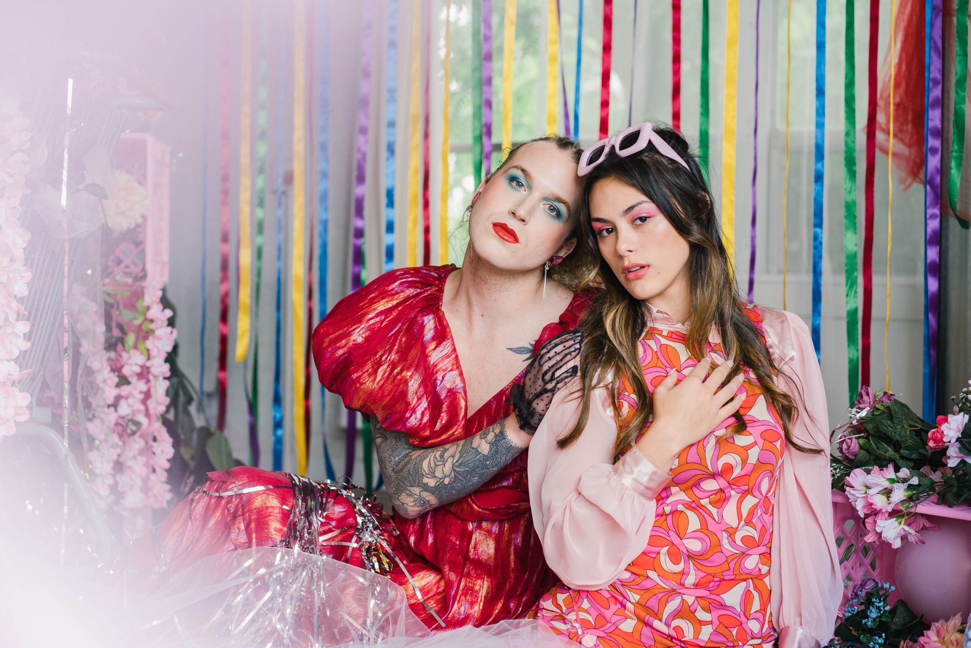 Two women are sitting next to each other in front of a colorful background for a fashion photo shoot.