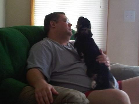 Man seated, holding a small black dog. Dog stands on hind legs, looking up at the man. Green chair, indoor setting.