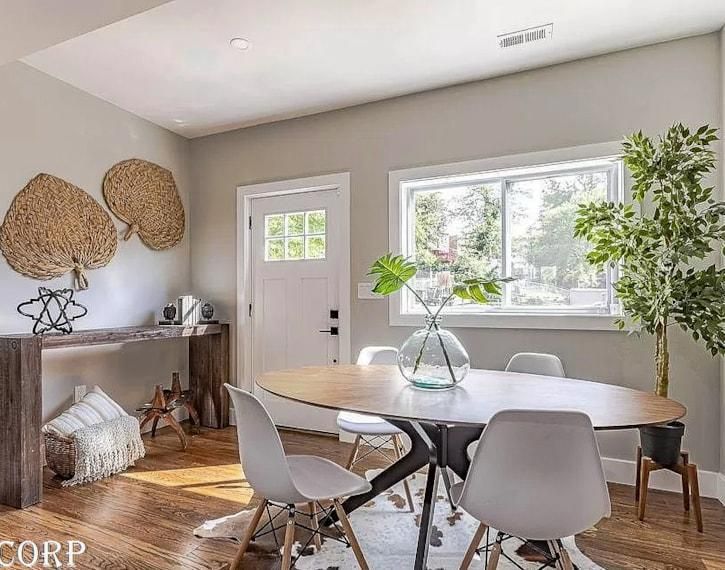 Bright dining room with oval table, white chairs, large window, woven wall decor, and potted greenery