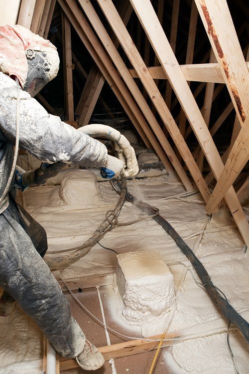 A man is spraying insulation on the floor of an attic.