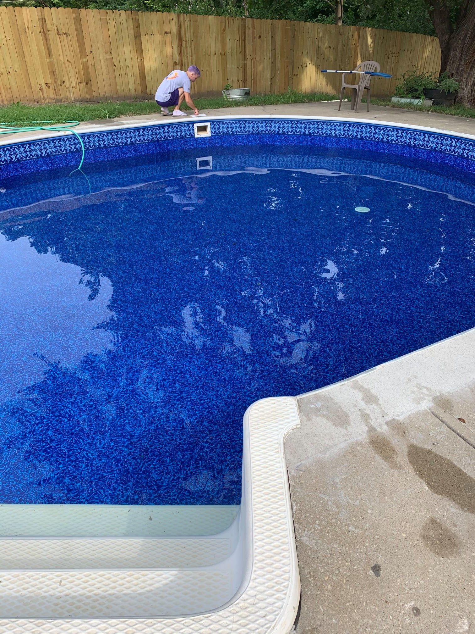Person cleaning a blue tile-lined pool. Concrete deck with steps in foreground, wooden fence in background.