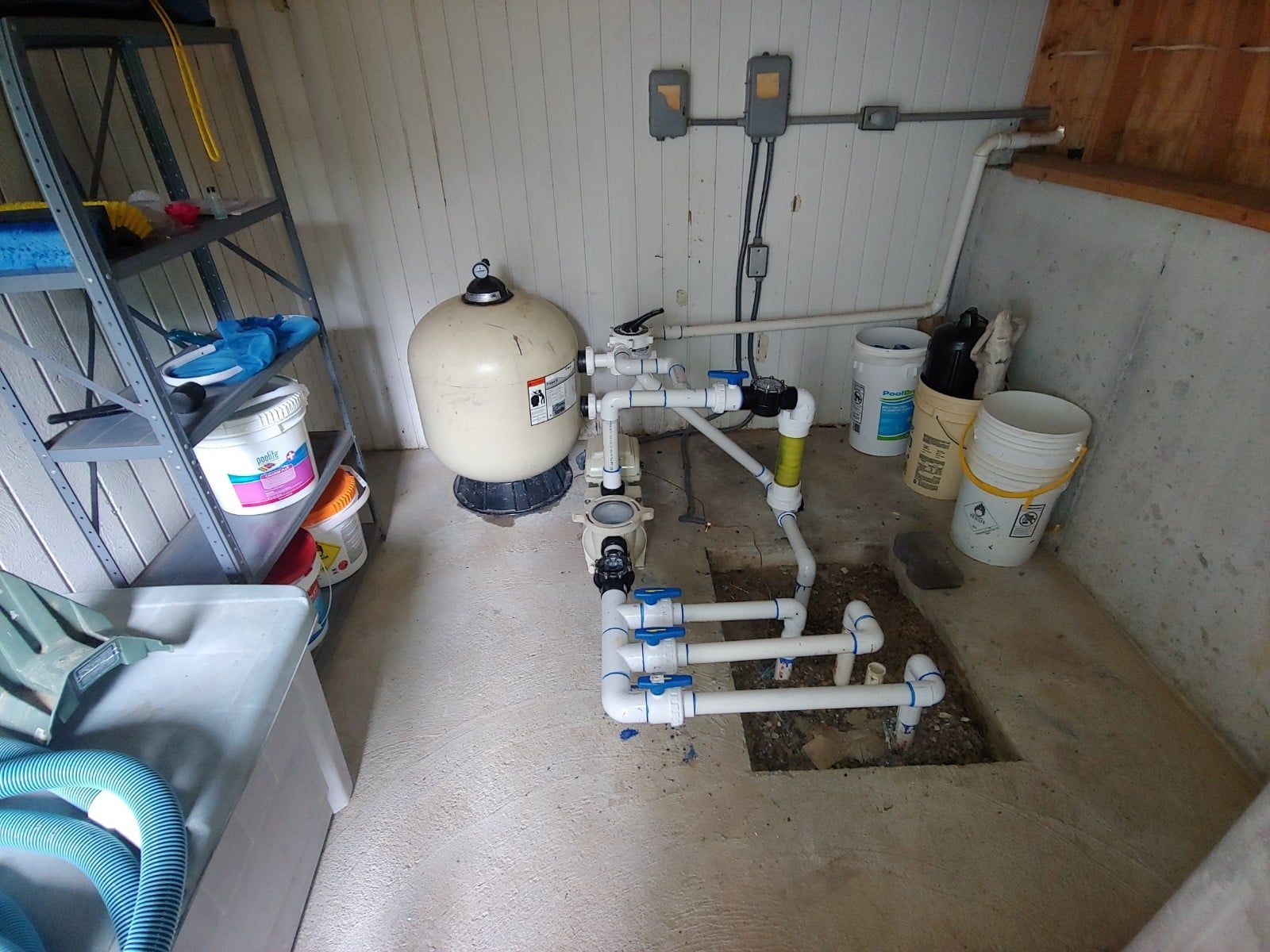 Pool pump and filter system in a utility room; white pipes, beige filter, concrete floor, and tools.