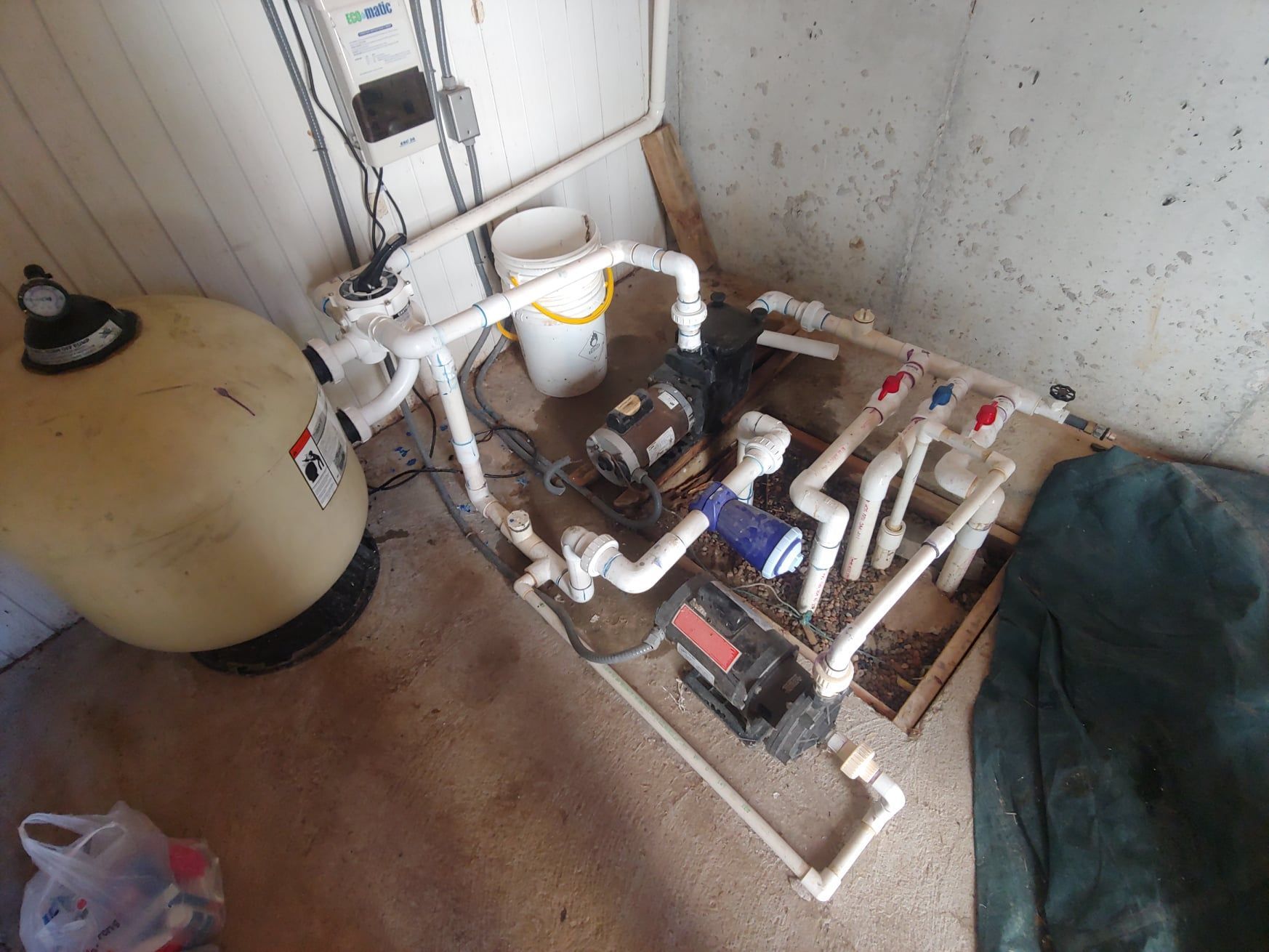 Pool equipment: Filter, pump, and plumbing in a utility room with a green tarp and bucket.