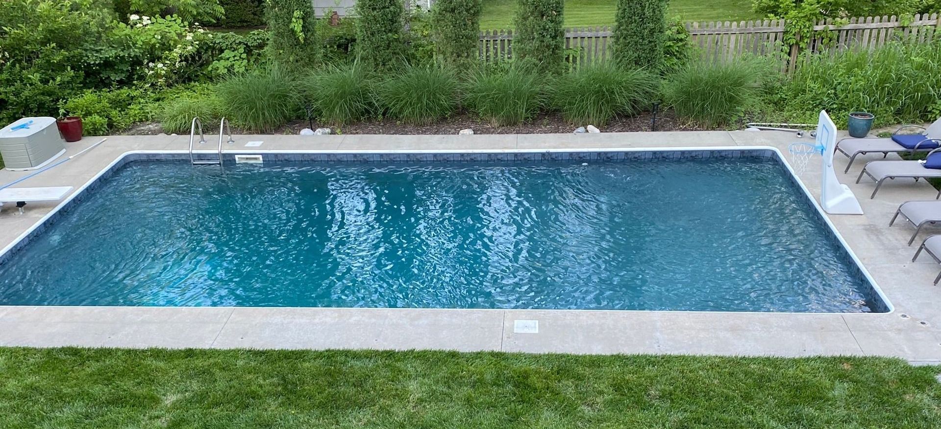 Rectangular swimming pool with clear blue water surrounded by concrete and green grass.