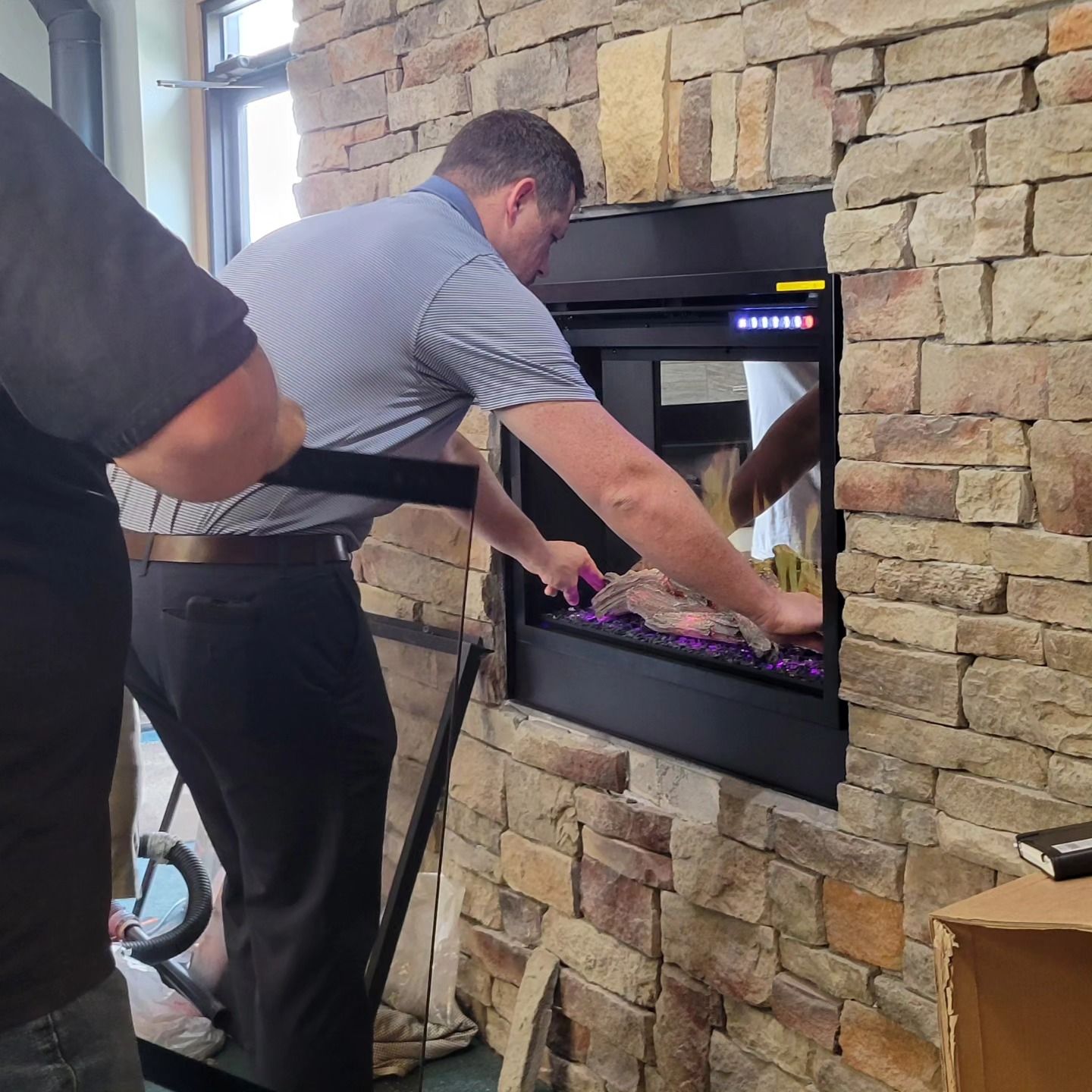 Two men installing a black electric fireplace in a stone wall. One adjusts the decorative rocks.