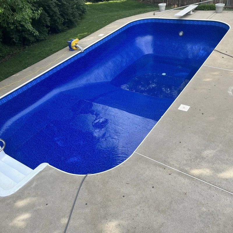Blue-tiled empty swimming pool, surrounded by concrete, steps in the corner, and green grass.