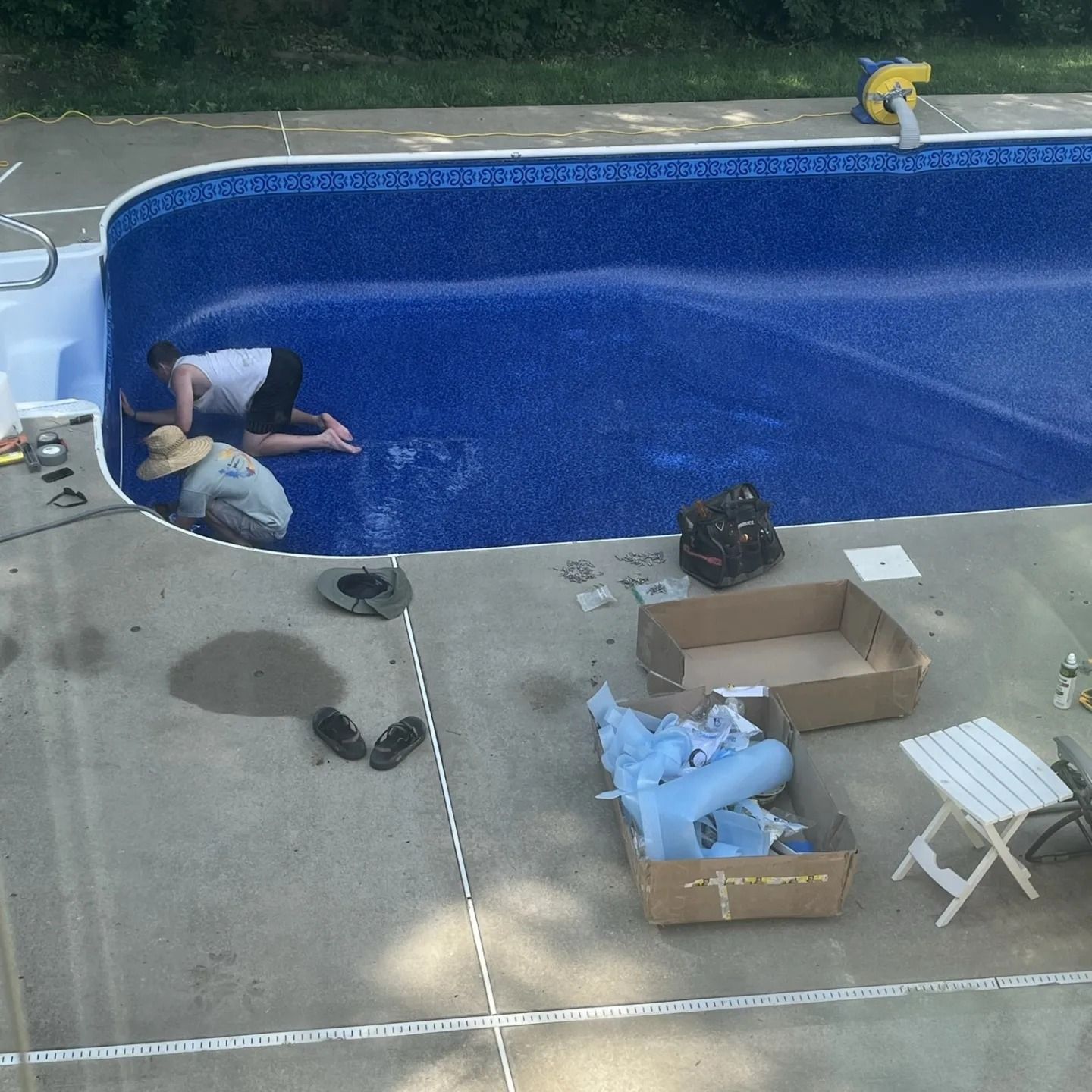 Two people installing a blue pool liner in an outdoor pool. Boxes and tools are nearby on concrete.