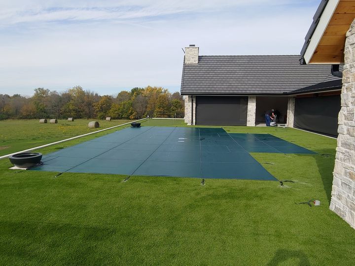 Pool covered with blue tarp, next to a house with green grass, and black garage doors.