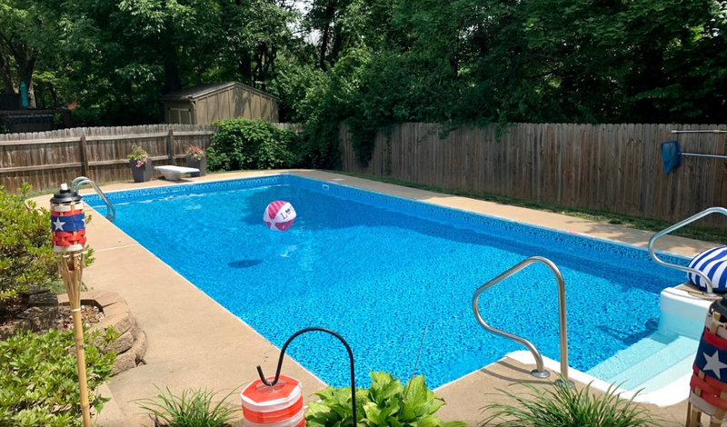 Rectangular backyard pool with blue water and a colorful beach ball. Surrounded by a concrete deck, fence, and greenery.