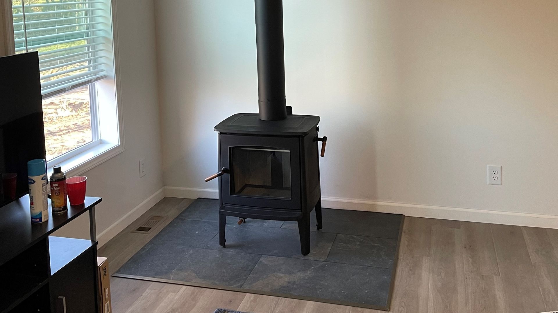 Black wood-burning stove in a room with a window, a TV stand, and a gray hearth pad.
