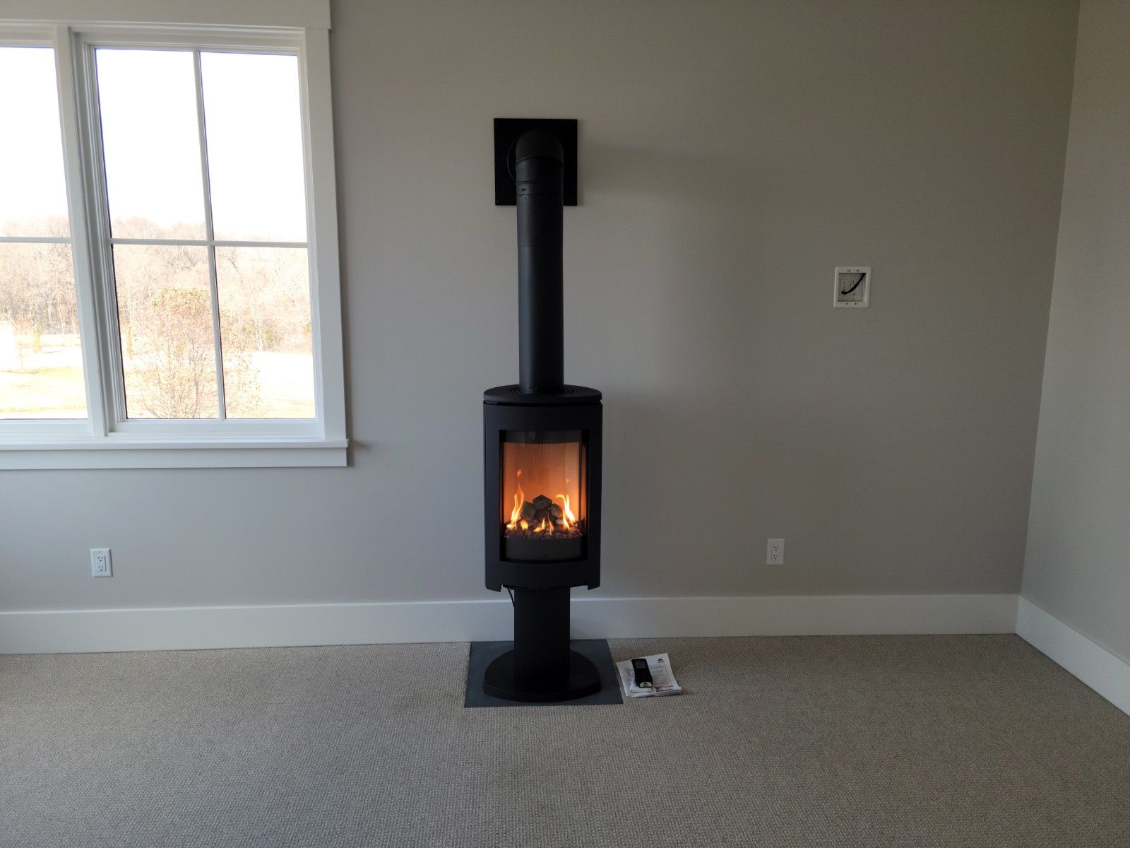 A black wood-burning stove with fire in a gray-walled room, next to a window on carpet.