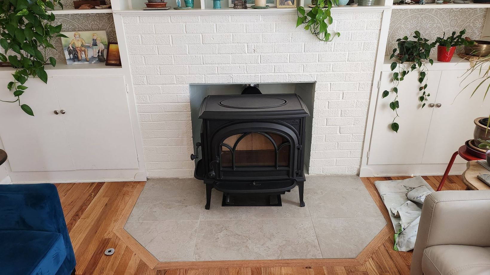 Black stove centered in a white brick fireplace. Wooden floor, shelves with plants, and a blue couch.