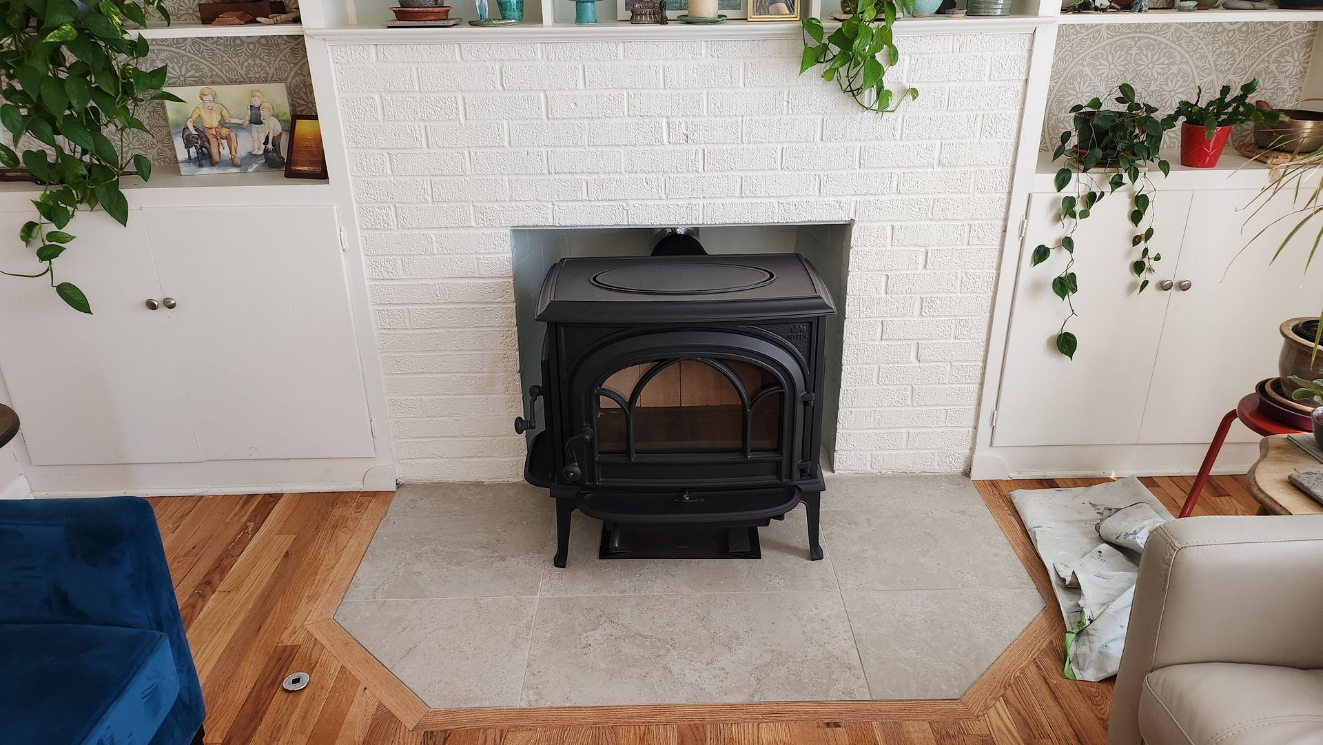 Black stove centered in a white brick fireplace. Wooden floor, shelves with plants, and a blue couch.