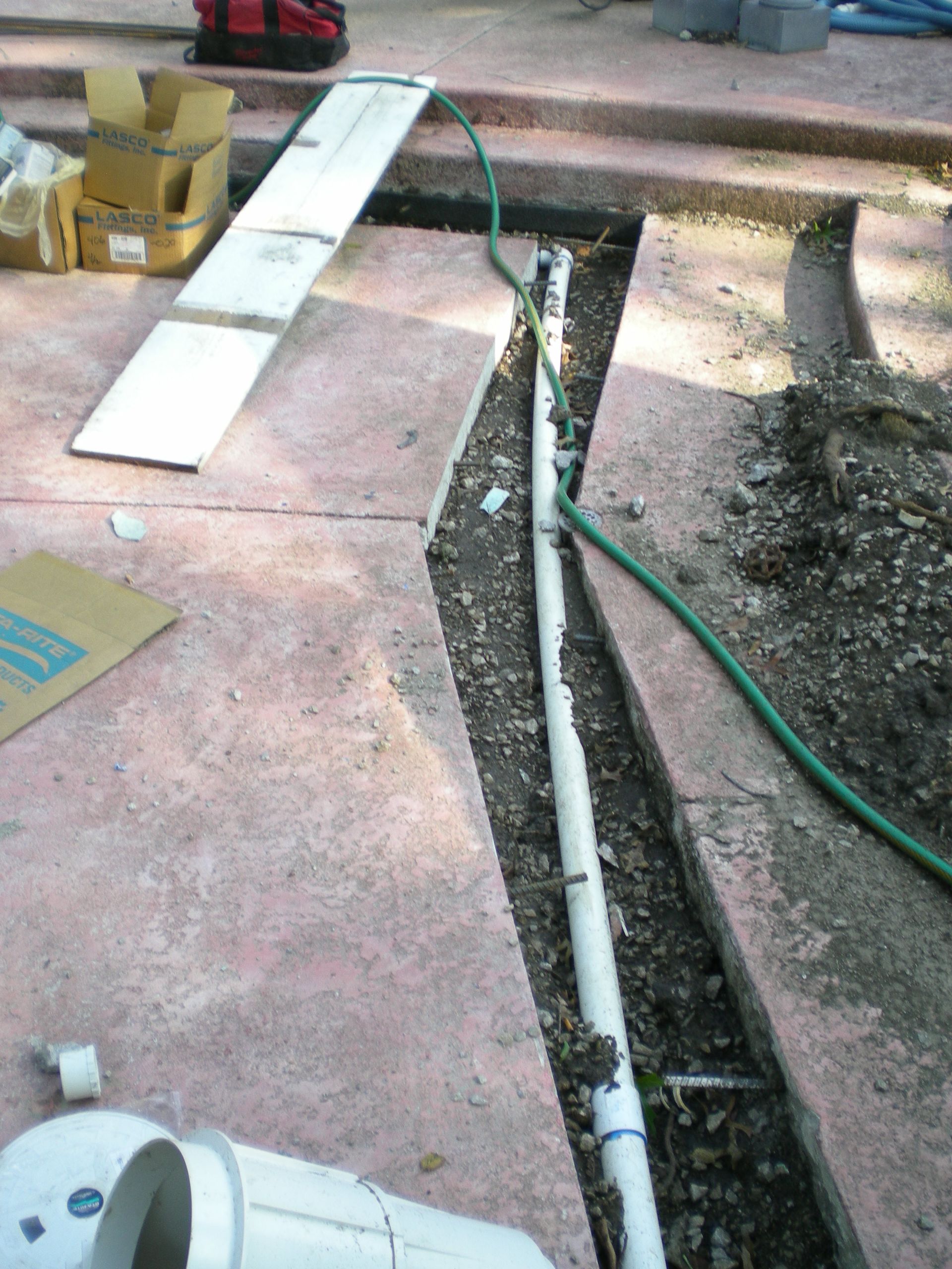 Trench in red concrete with white pipe inside; a green hose runs nearby. Tools and cardboard boxes are in the background.