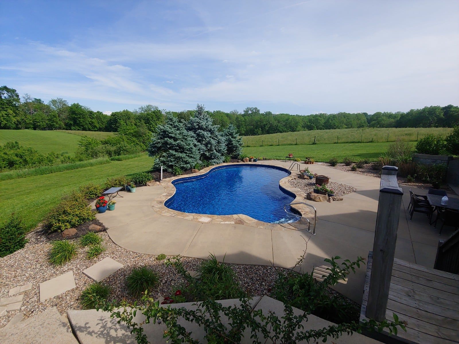 Pool surrounded by concrete patio, trees, and green fields under a blue sky.