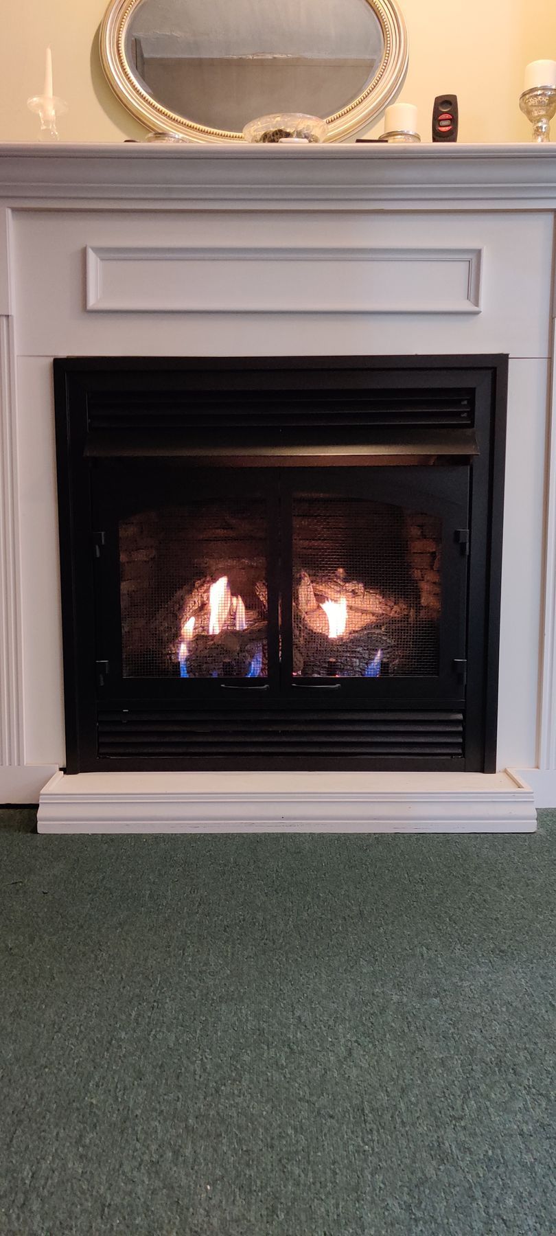 Fireplace with flames in a white mantle, mirror above, and green carpet.