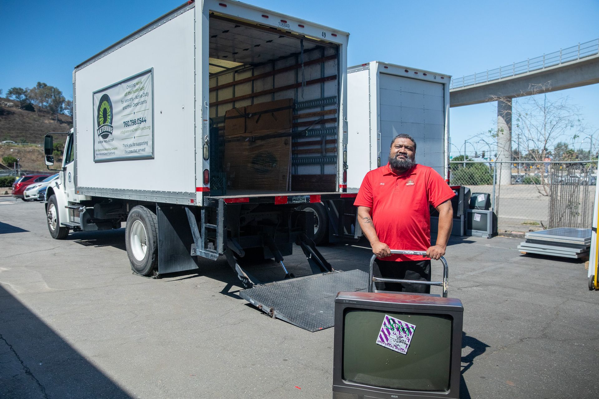 Man in red shirt pushes a dolly with a TV towards a moving truck with an open back ramp.