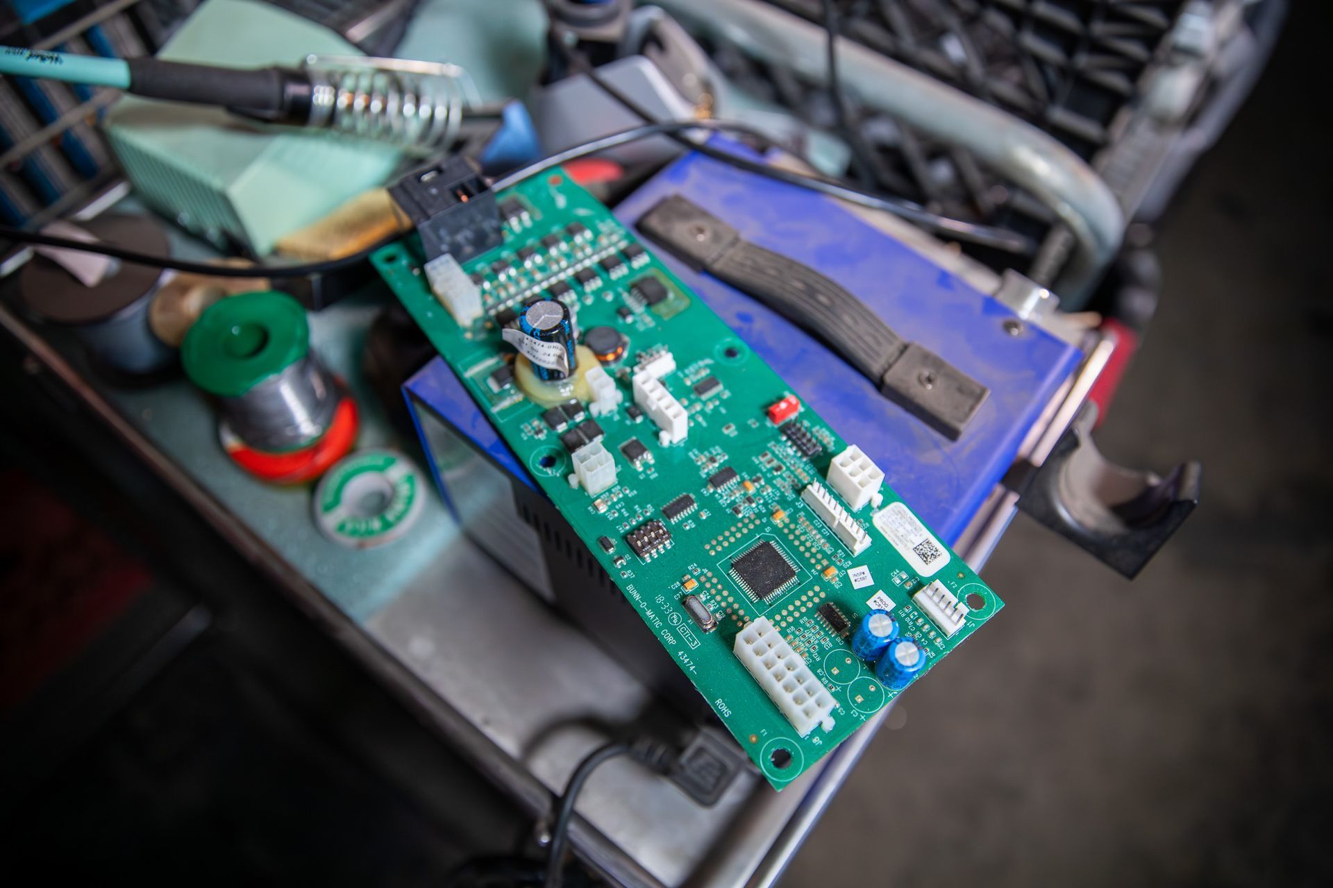 Green circuit board with electronic components, resting on a blue object, near a soldering iron and spools of solder.