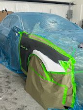 Car in a body shop, partially covered in blue plastic. A door and fender are painted black and white, masked with green tape  | Axe-A-Dent Auto Collision