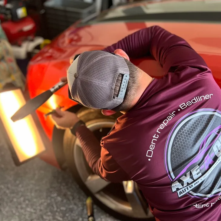 Man in red shirt repairing a red car, using tools. The car is near a tire.
