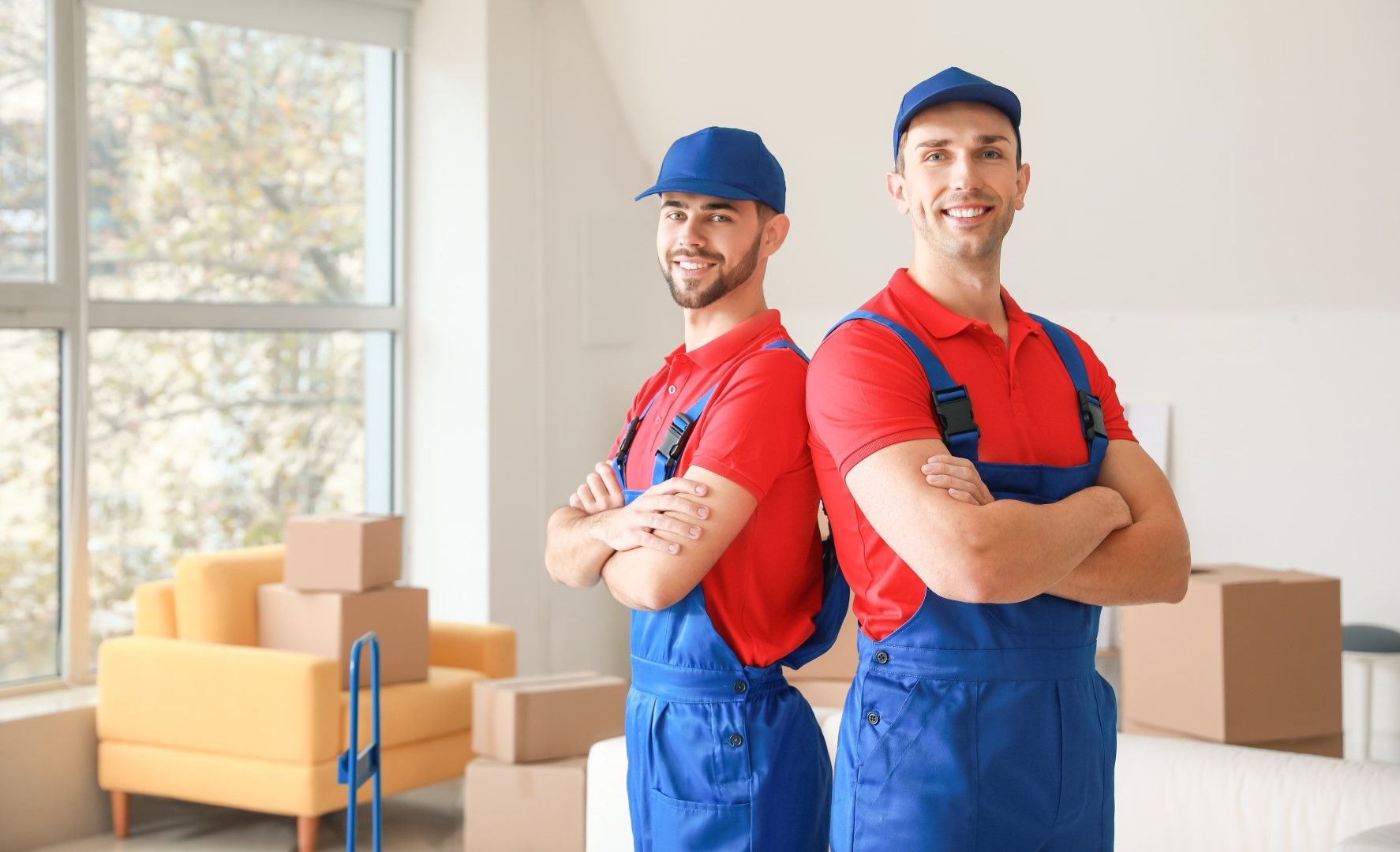 Two movers are standing back to back in a living room with their arms crossed.