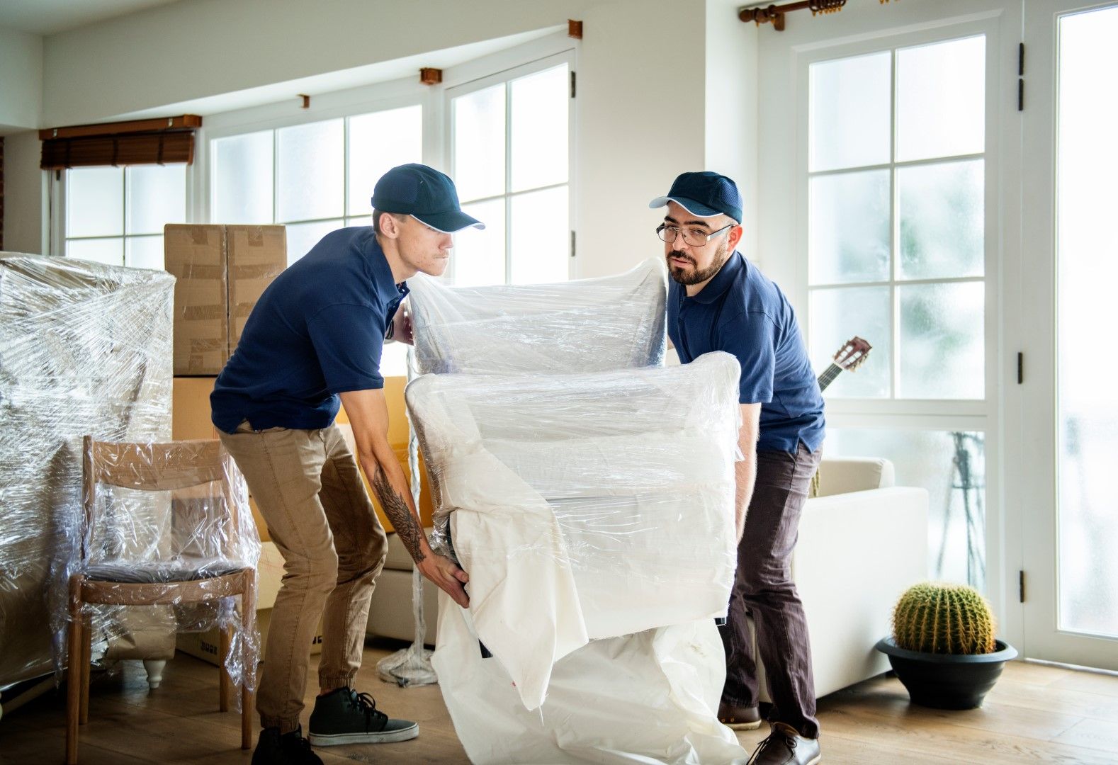 Two men are carrying a chair wrapped in plastic in a living room.