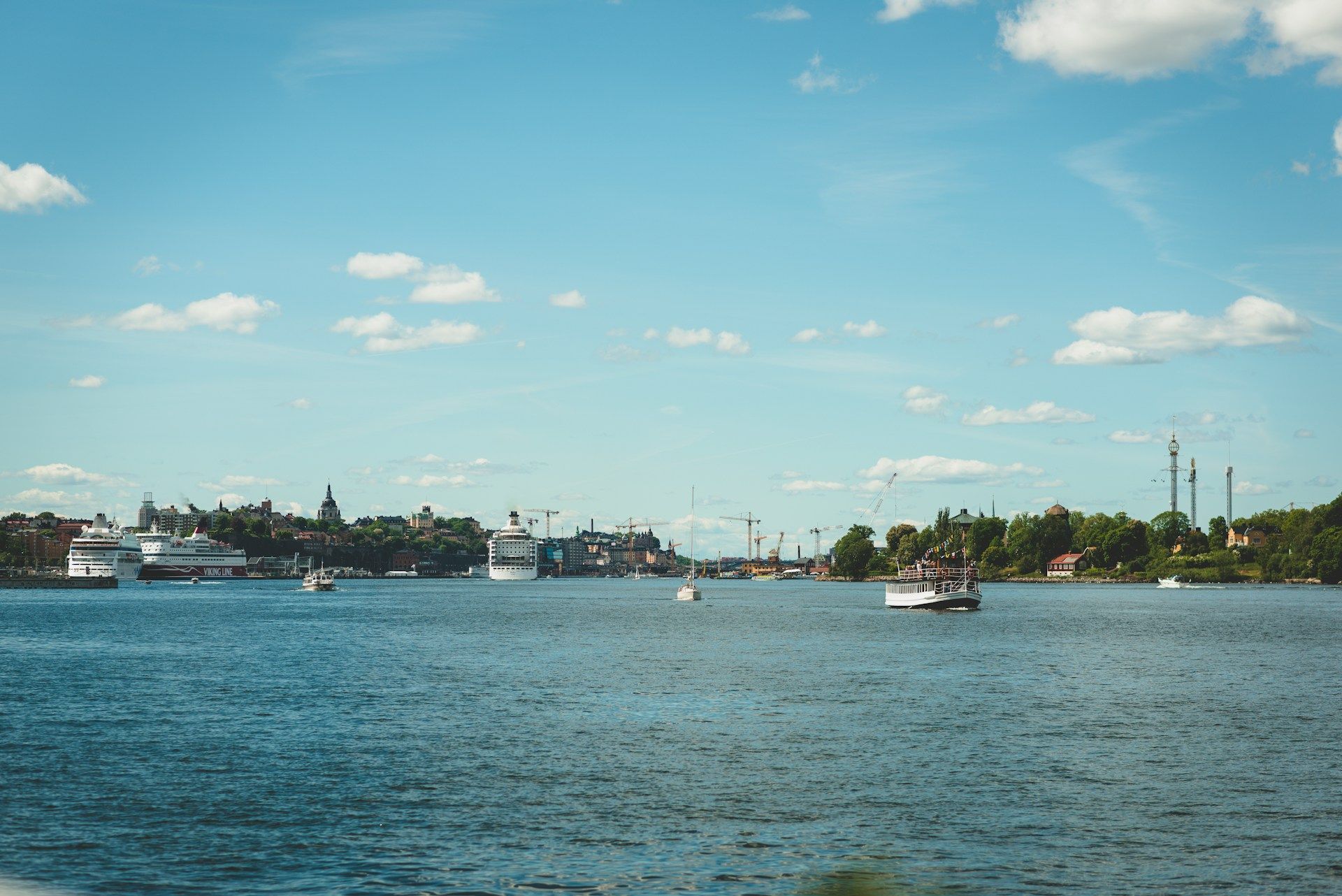 Boats and cruise ships navigate the Stockholm inlet surrounded by trees and distant city buildings.