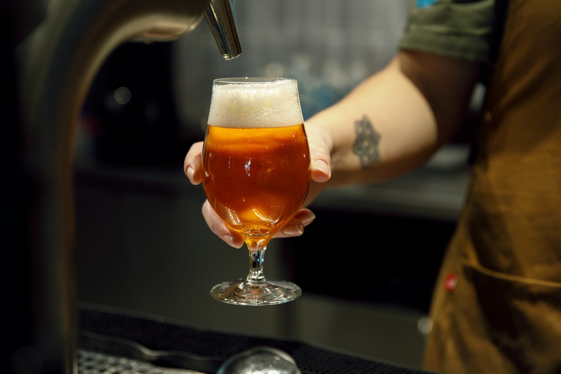 A hand holds a stemmed glass filled with amber beer under a beer tap.