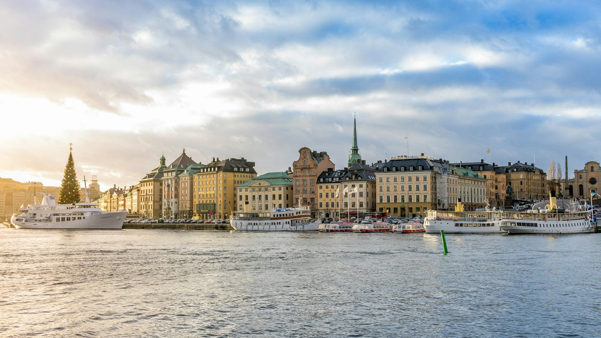 Stockholm's historic Gamla Stan waterfront with colorful buildings, a Christmas tree, and boats under a cloudy sky.