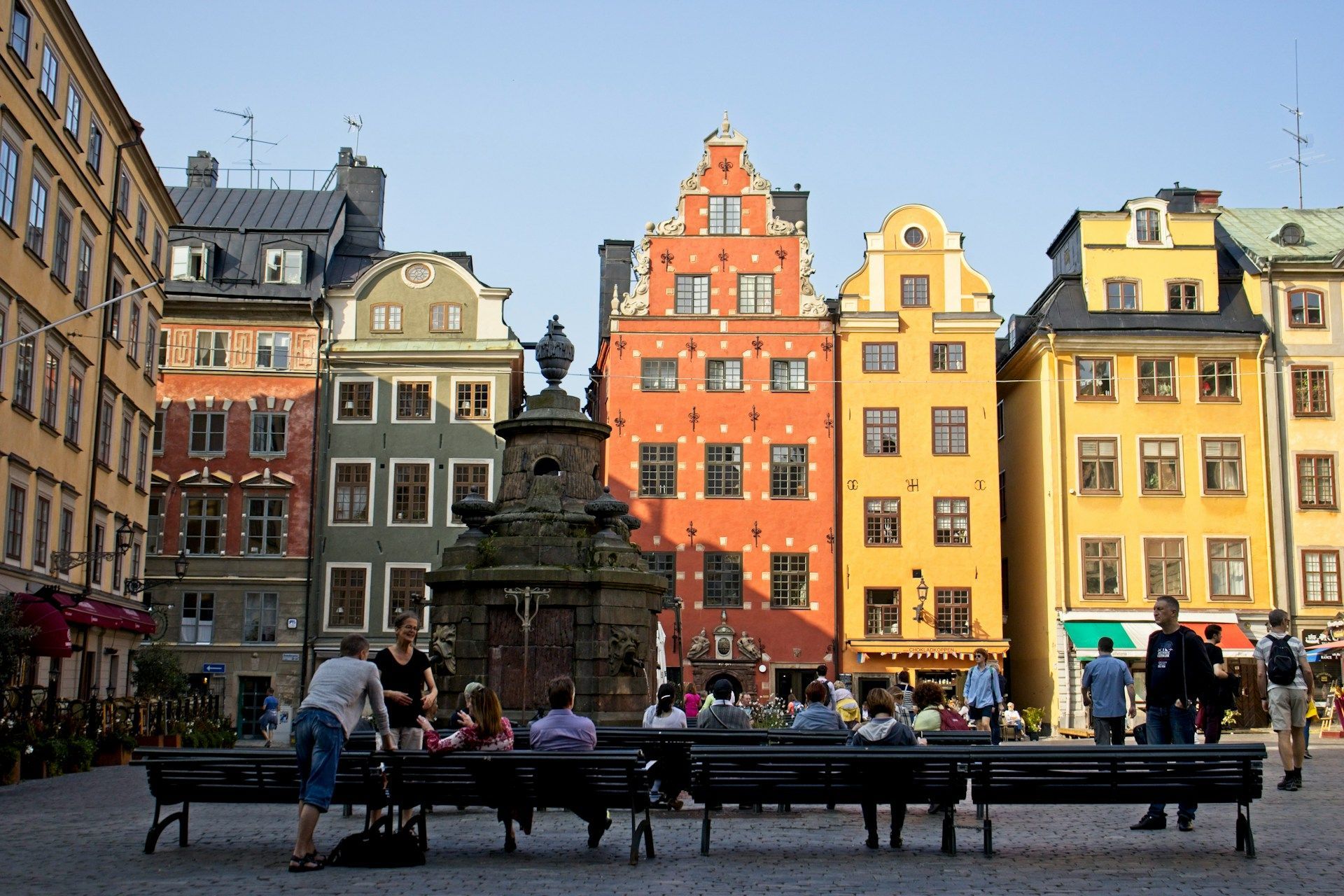 People sit on benches in Stortorget, a sunny town square surrounded by historic colorful buildings in Stockholm.